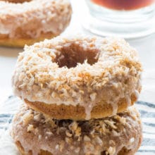 A stack of toasted coconut donuts sits on a kitchen towel with another donut in the background next to a cup of tea