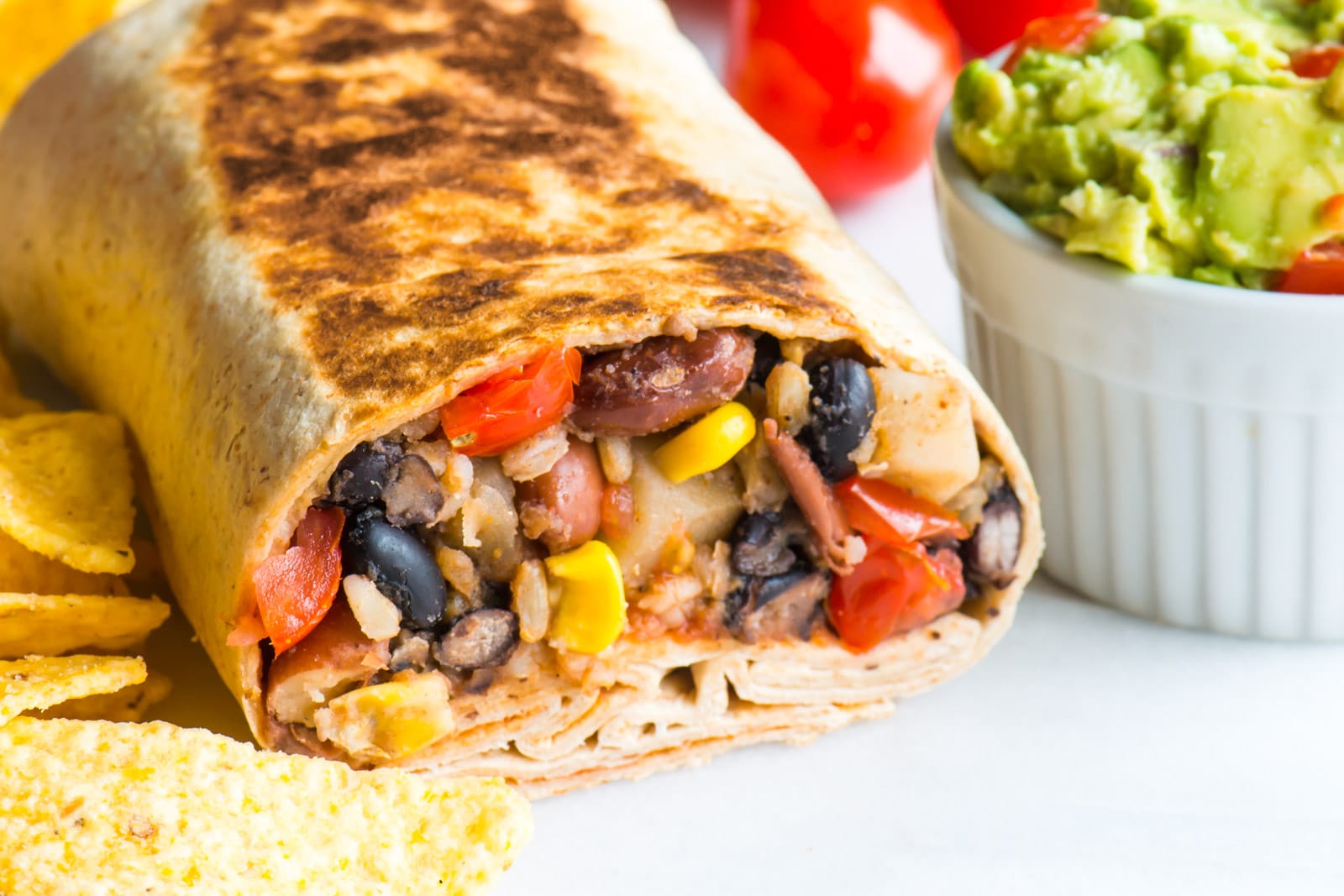 A vegan black bean burrito is cut in half to reveal beans and rice. It sits next to guacamole, fresh cherry tomatoes, and tortilla chips.