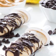 Two banana popsicles drizzled in chocolate sit in front of bananas and more ingredients.