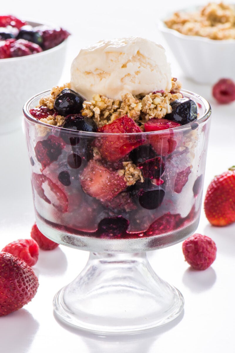 A bowl of vegan no-bake cherry fruit crisp in a serving dish with ice cream on top and other toppings behind it.
