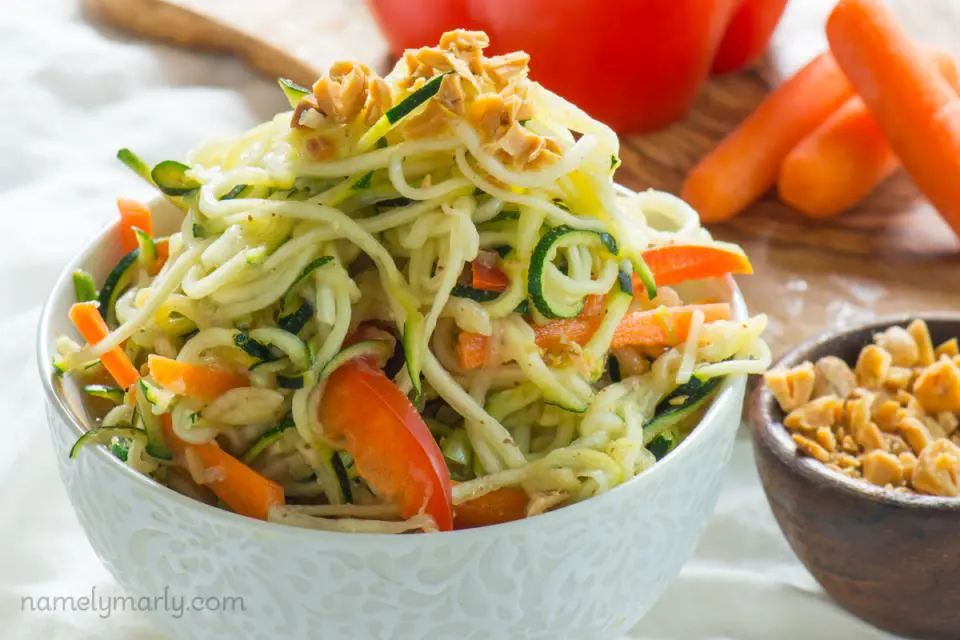 A bowl of zucchini noodles has chopped peanuts on top. There's a wooden bowl with more chopped peanuts on the side and a cutting board with veggies in the background.