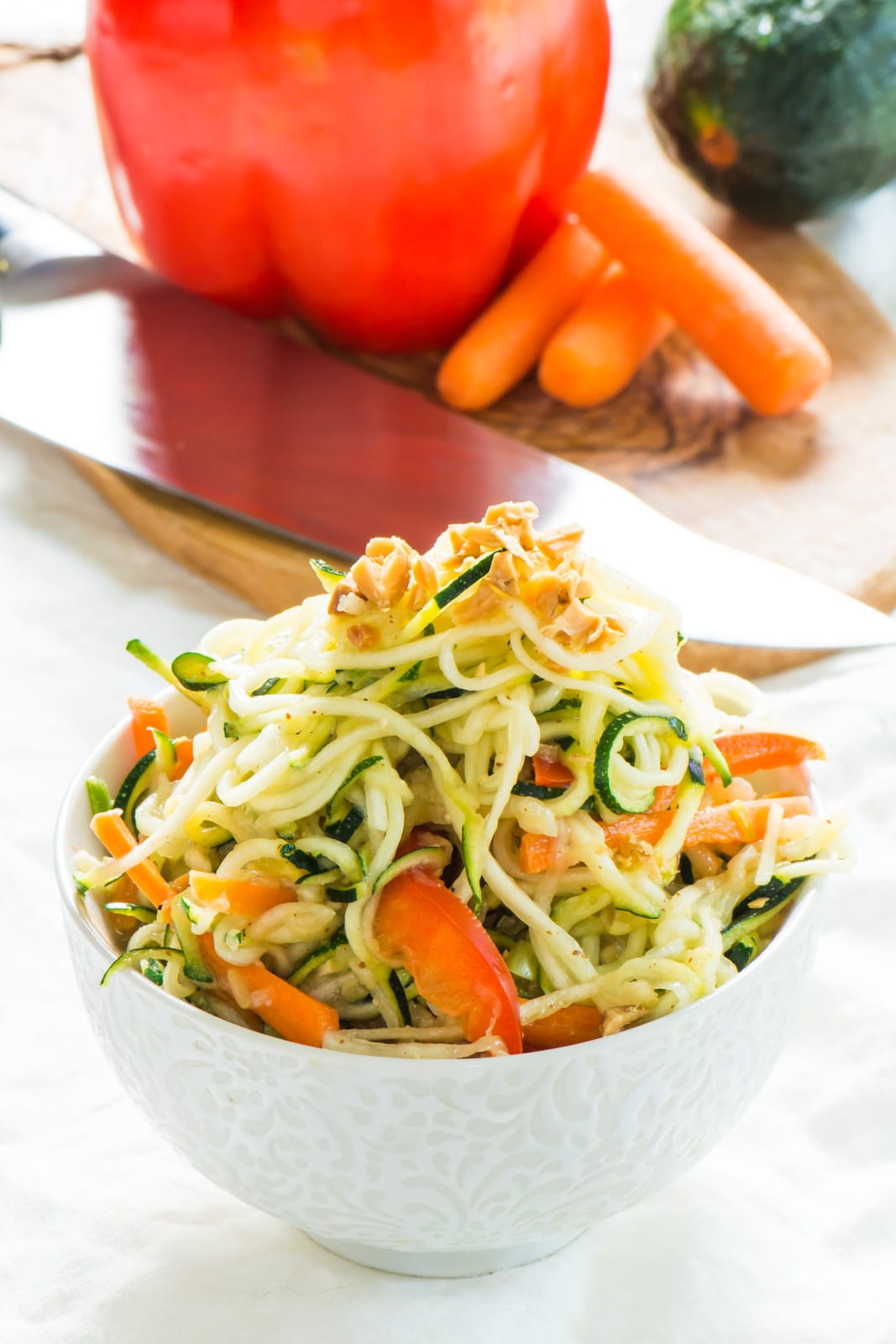Pad Thai Zucchini Noodles are in a bowl with chopped peanuts on top. There's a cutting board in the background with a knife, carrots, a red bell pepper, and a zucchini.