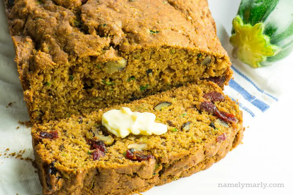 A loaf of zucchini bread has a slice cut out, sitting next to a zucchini. There's a pat of butter on the slice.