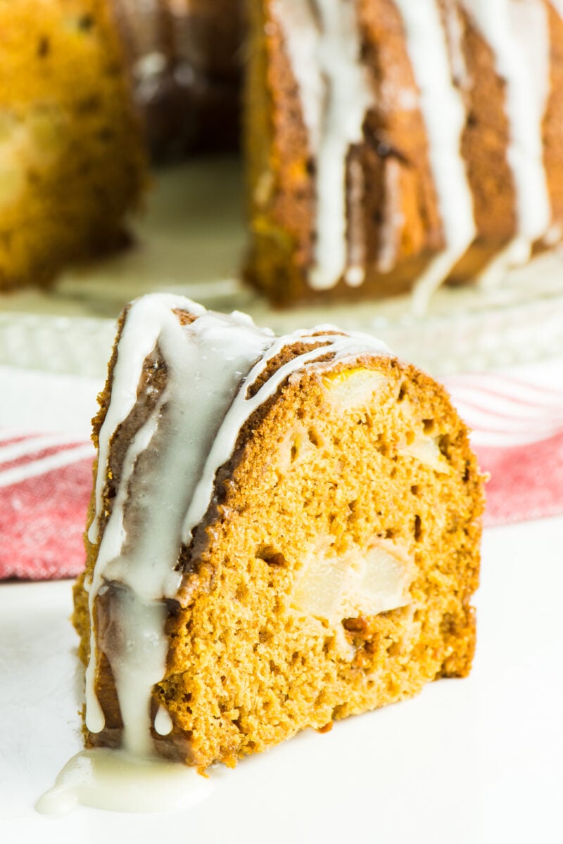A slice of fresh pear cake sits on a plate in front of the rest of the cake.