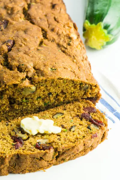 A loaf of vegan healthy zucchini bread with a slice cut off sits next to a zucchini.