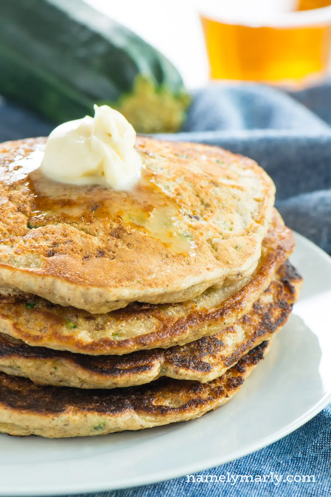 A stack of pancakes has melted butter on top. A bowl behind it holds maple syrup. A zucchini sits behind it too.