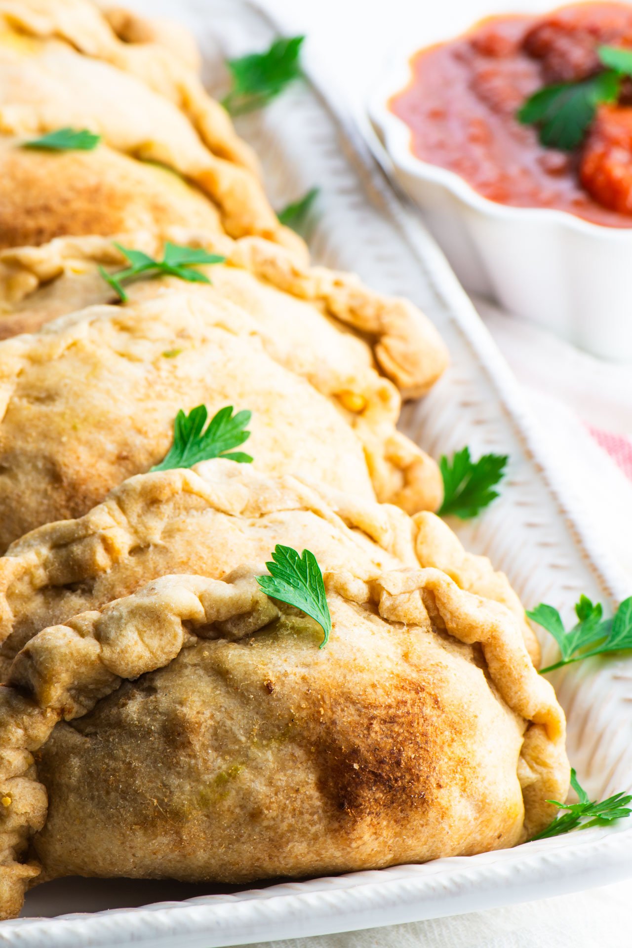 A row of vegan beef empanadas on a serving platter next to a bowl of red sauce.