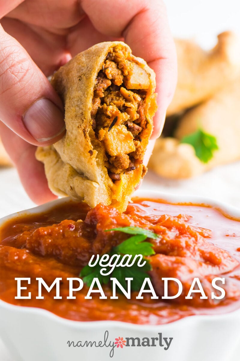 A hand holds an empanada about to dip into a bowl full of marinara sauce with text that reads Vegan Empanadas