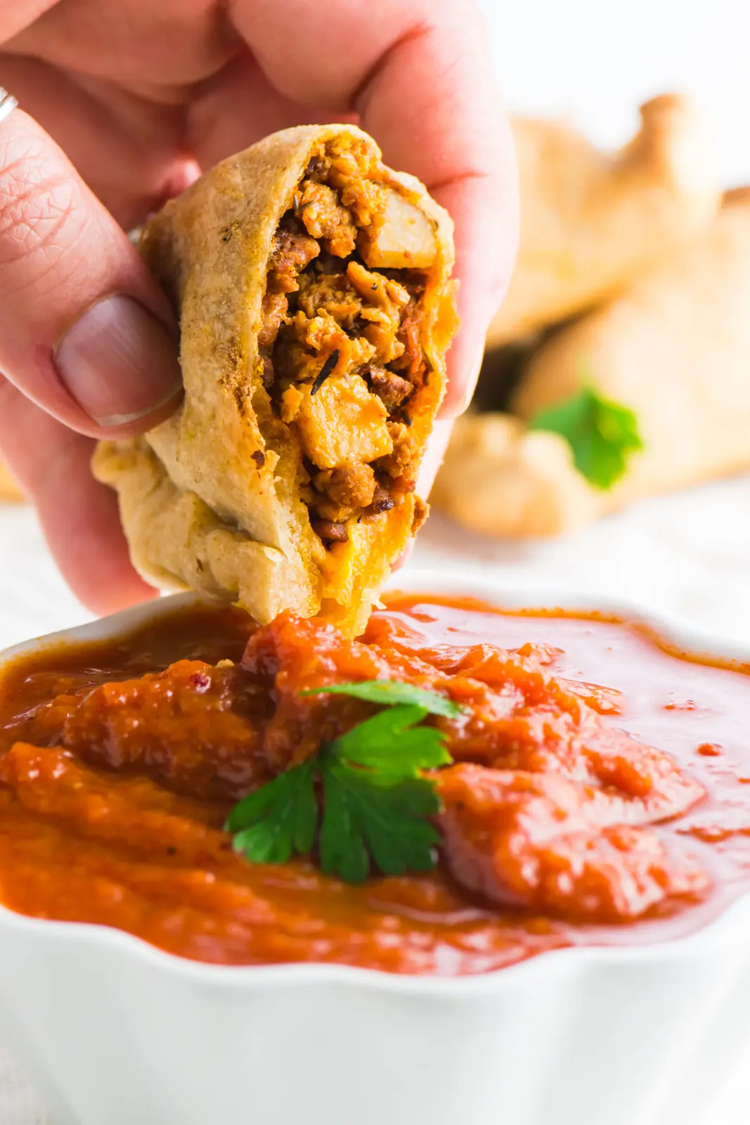 A hand holds a vegan empanada, dipping it into a red sauce.