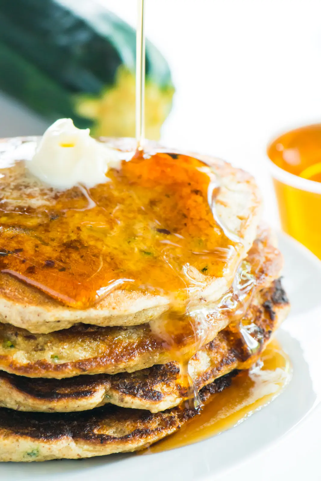 A stack of zucchini pancakes has syrup drizzling over the top of it. A bowl of syrup and a zucchini sits behind it.