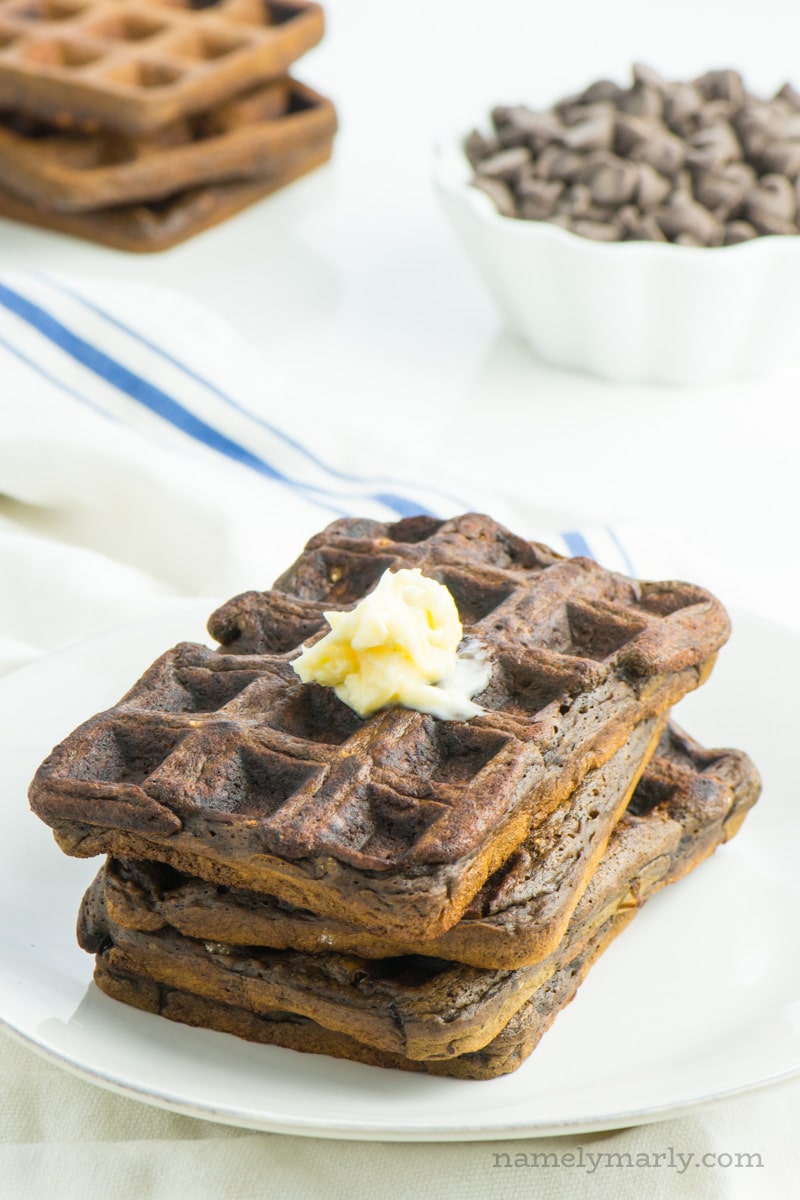 A stack of chocolate waffles with melted butter on top and a bowl of chocolate chips behind it.