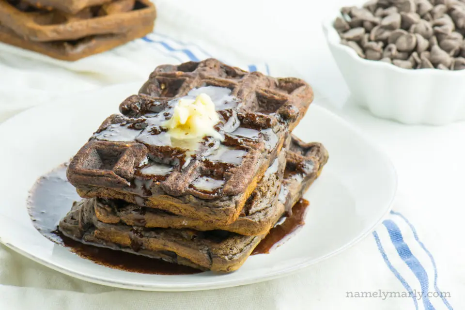 A stack of vegan waffles on a plate with chocolate syrup. A bowl of chocolate chips and more waffles behind it.