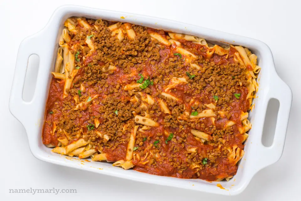 Looking down on a casserole dish full of this cooked penne topped with red sauce and veggie crumbles.