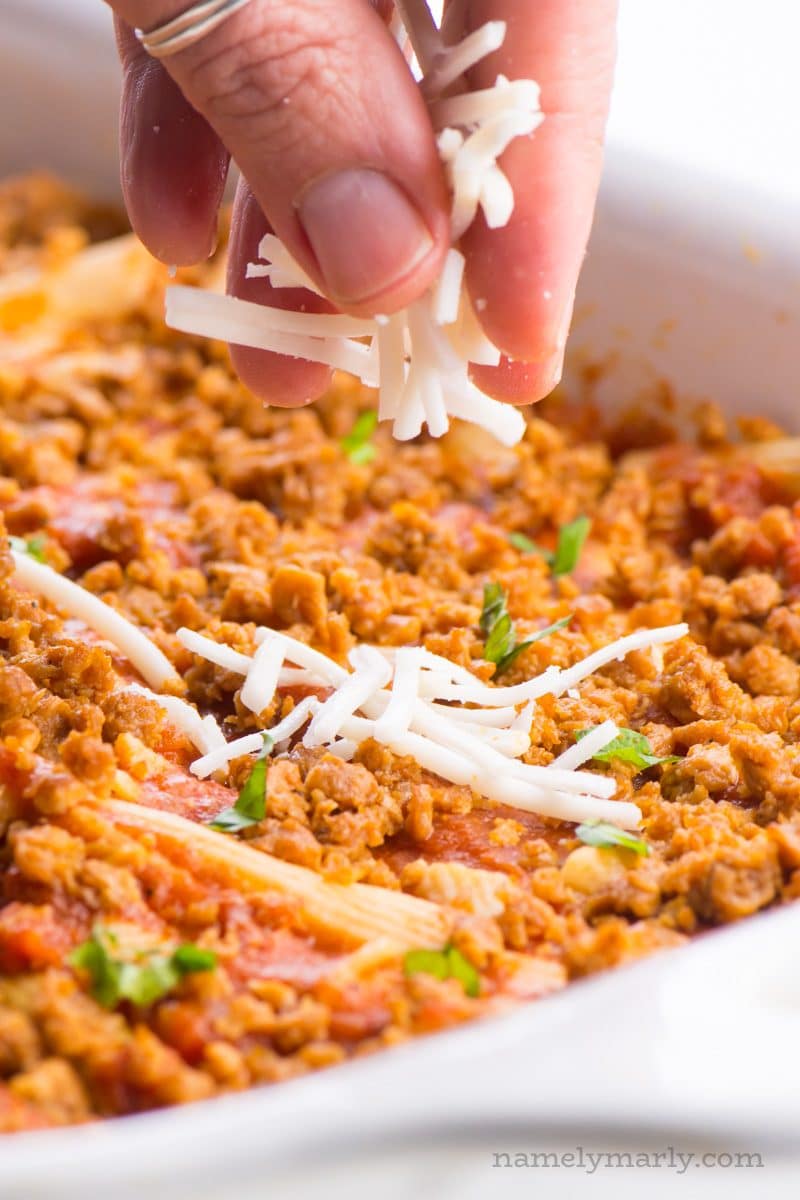 A hand is spreading vegan mozzarella over a casserole dish.