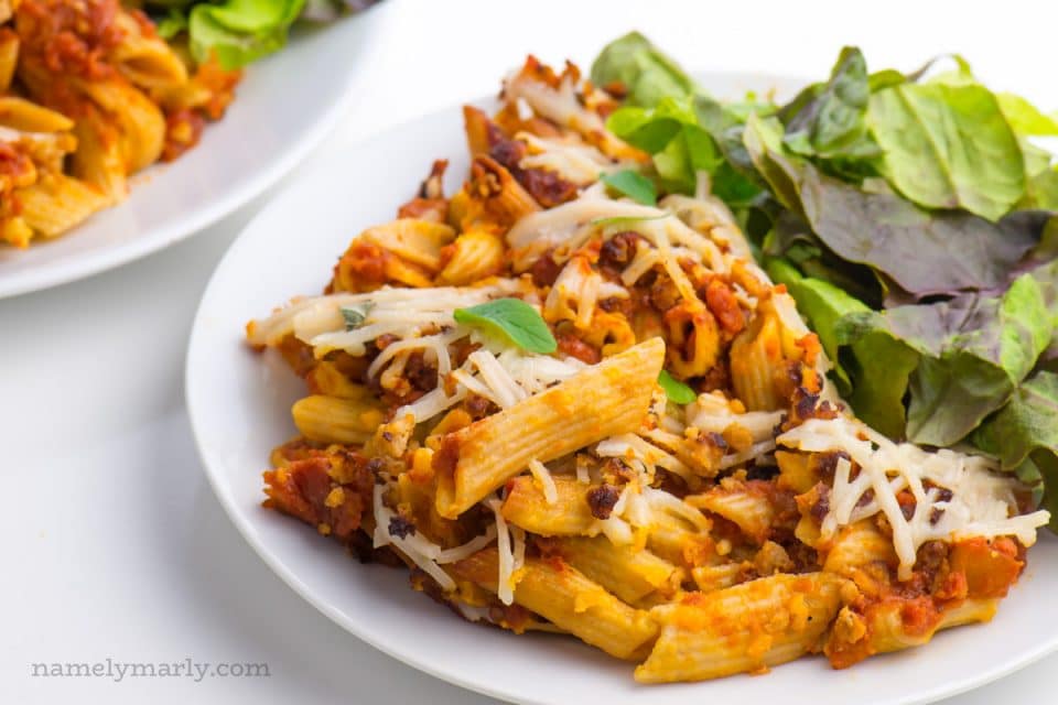 A plate of vegan pasta bake and a green leafy salad sits beside another plate full of pasta as well.