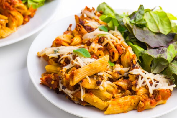 A plate of vegan pasta bake and a green leafy salad sits beside another plate full of pasta as well.