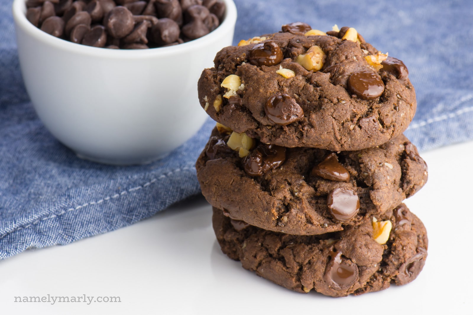 A stack of three chocolate walnut cookies sits on a white surface with a blue napkin folded behind