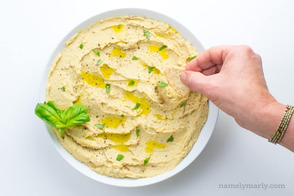 A. hand holds a cracker, reaching into a bowl of onion hummus. There are fresh herbs on top of the. hummus.