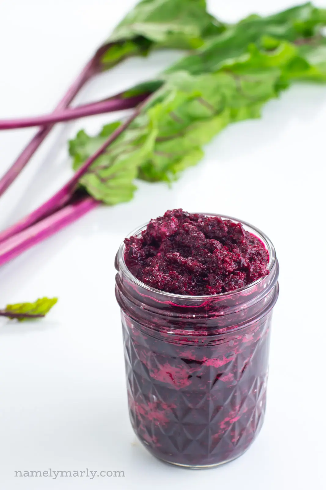 A jar of bright red mashed beets sits next to beet greens with bright pink stems.
