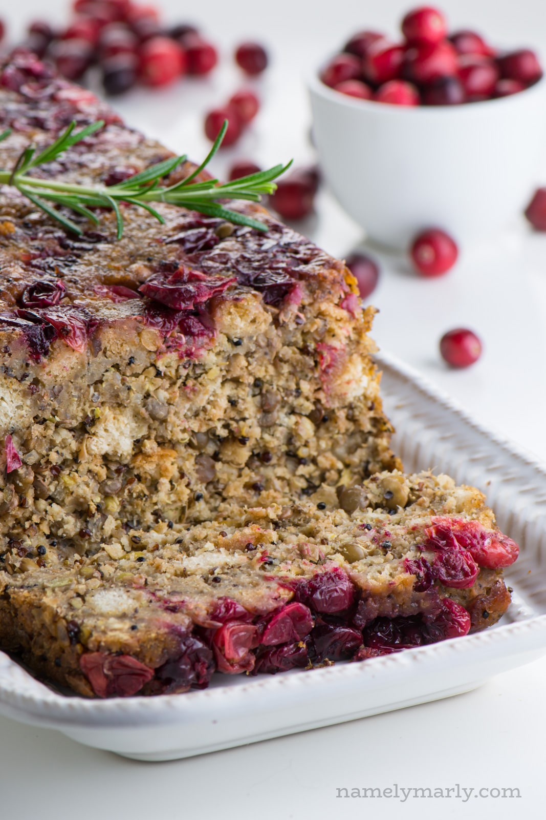 A slice of Vegan Lentil Loaf on a plate in front of the remaining loaf with a small bowl of cranberries in the back.