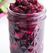 A jar holds pureed beets with beet greens behind it, with bright pink stems.