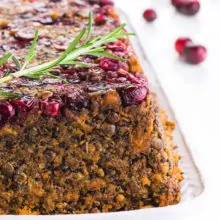 A cranberry-encrusted Lentil Loaf sits on a white plate with cranberries in the background.