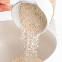 A hand tipping a measuring cup full of flour into a large bowl