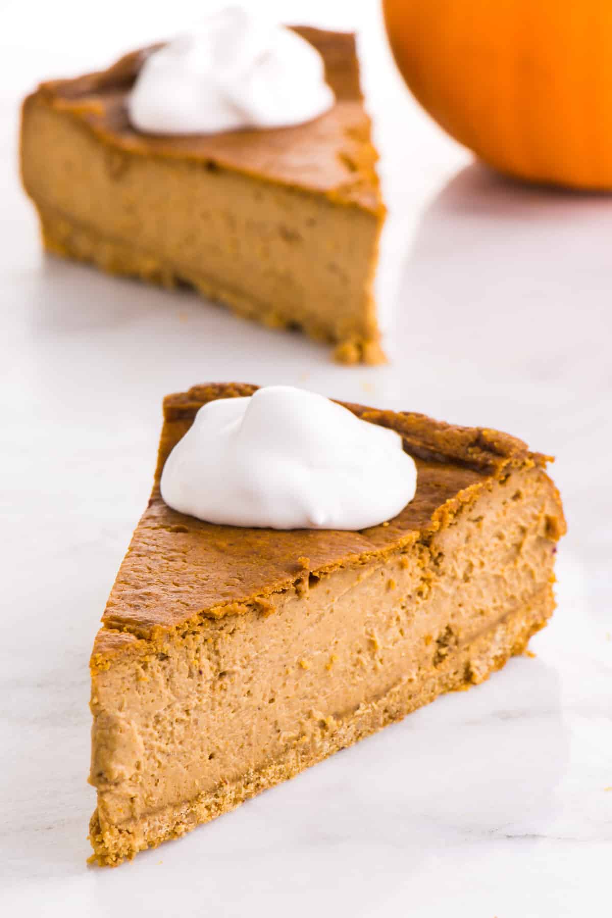 Two slices of vegan pumpkin cheesecake have whipped cream on top and sit next to  a small pumpkin.