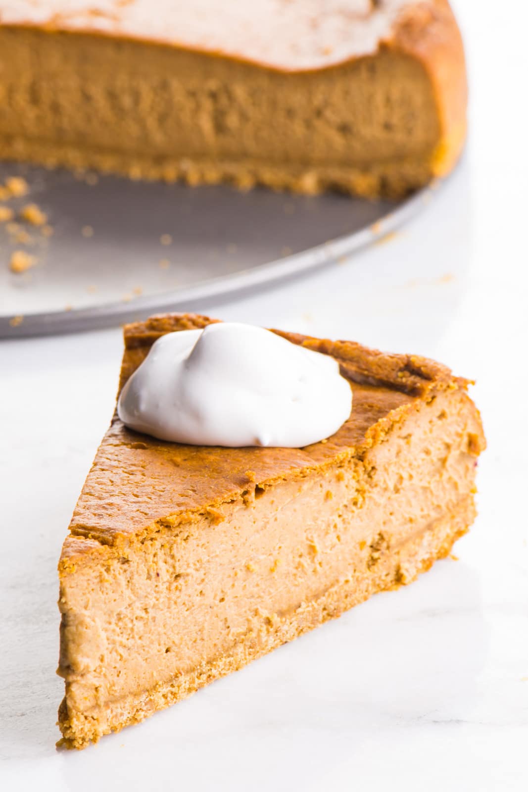 A slice of vegan pumpkin cheesecake is on a plate with vegan whipped cream on top. The rest of the cheesecake is behind it.