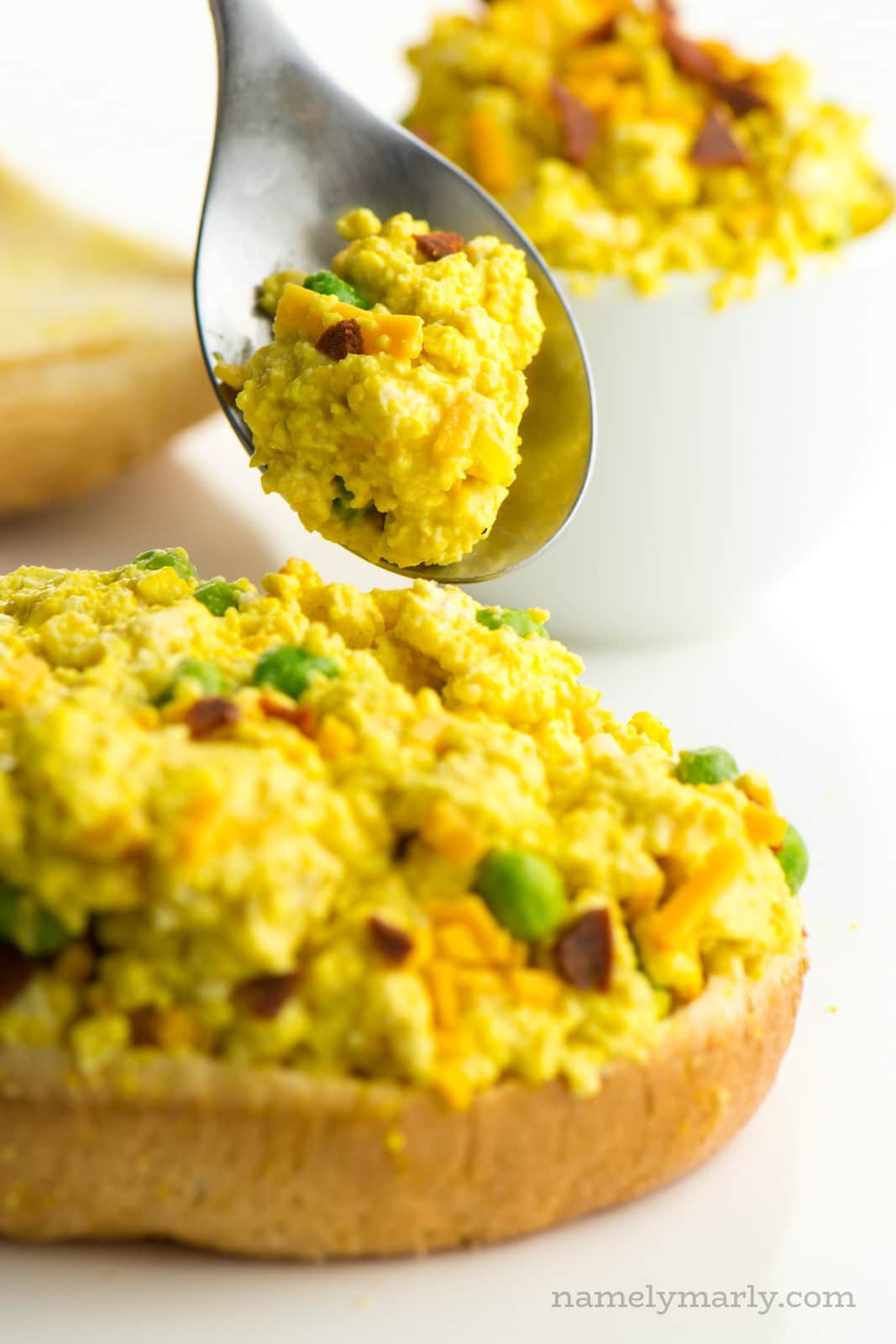 A spoon with vegan egg salad hovers over a sandwich loaded with the topping. A bowl with more of the ingredients is behind it.