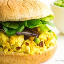 A bun has vegan egg salad along with greens. A bowl of frozen peas is in the background.