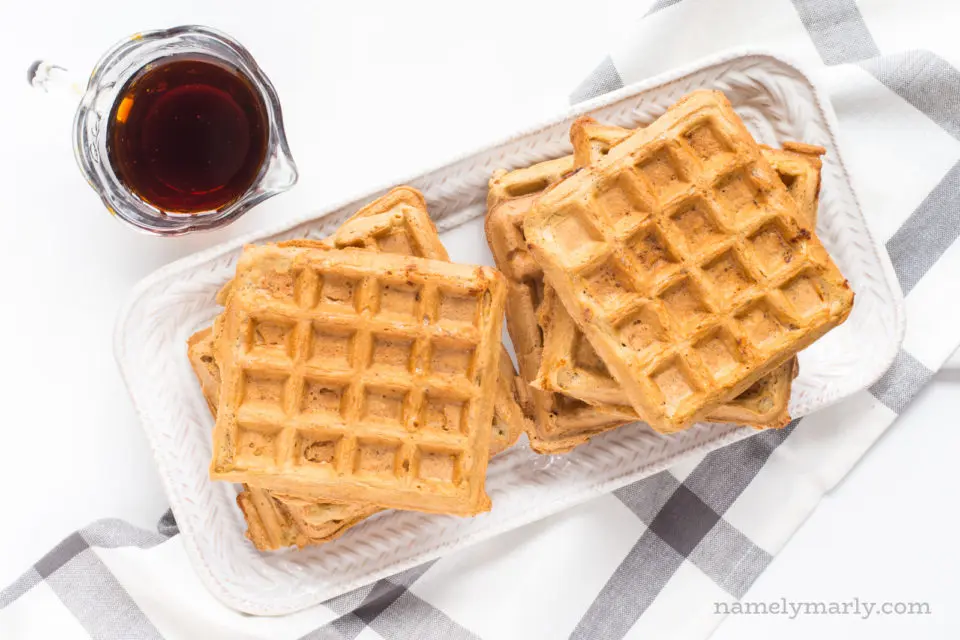 Vegan waffles sit stacked on a platter, with a dish towel underneath the platter. There is a small glass pitcher full of maple syrup be the waffles.