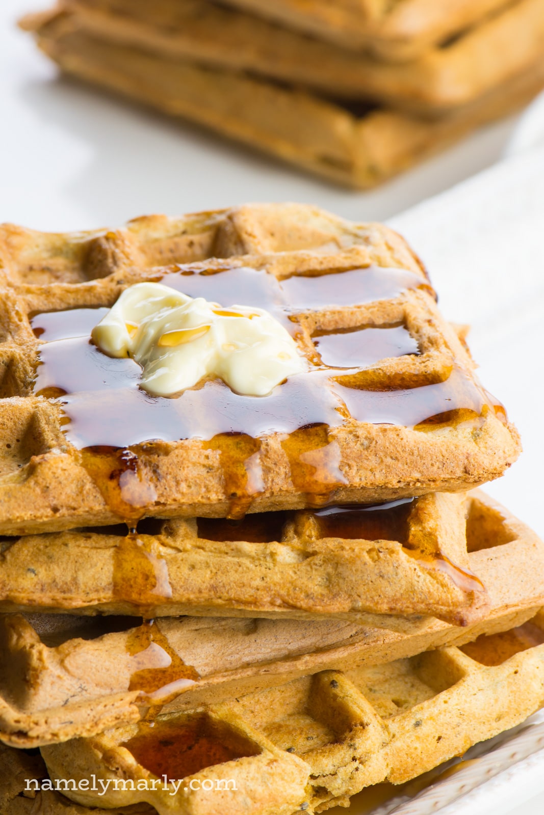 A stack of four whole wheat vegan waffles sit on a plate. There's melted vegan butter on the top waffle and it has maple syrup pooled in the waffle impressions on the top and oozing over the side. There are additional waffles in the background.