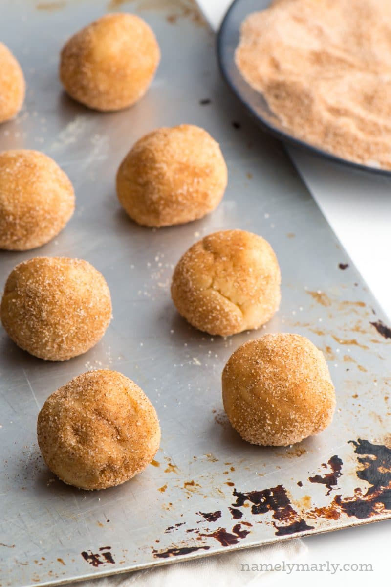 Snickerdoodle cookie dough balls are lined up a cookie baking sheet, waiting to to in the oven. A plate of cinnamon sugar sits nearby.
