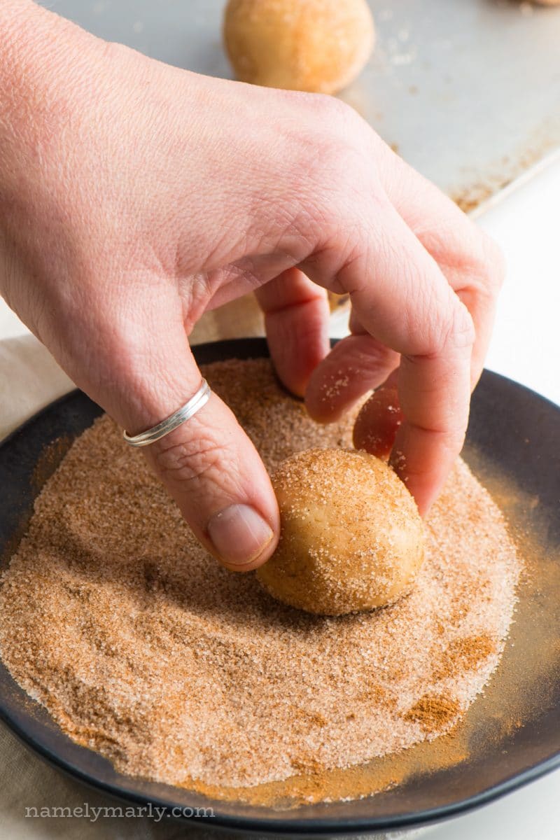 A hand is rolling a vegan snickerdoodle cookie dough ball in a plate full of cinnamon sugar mixed together.