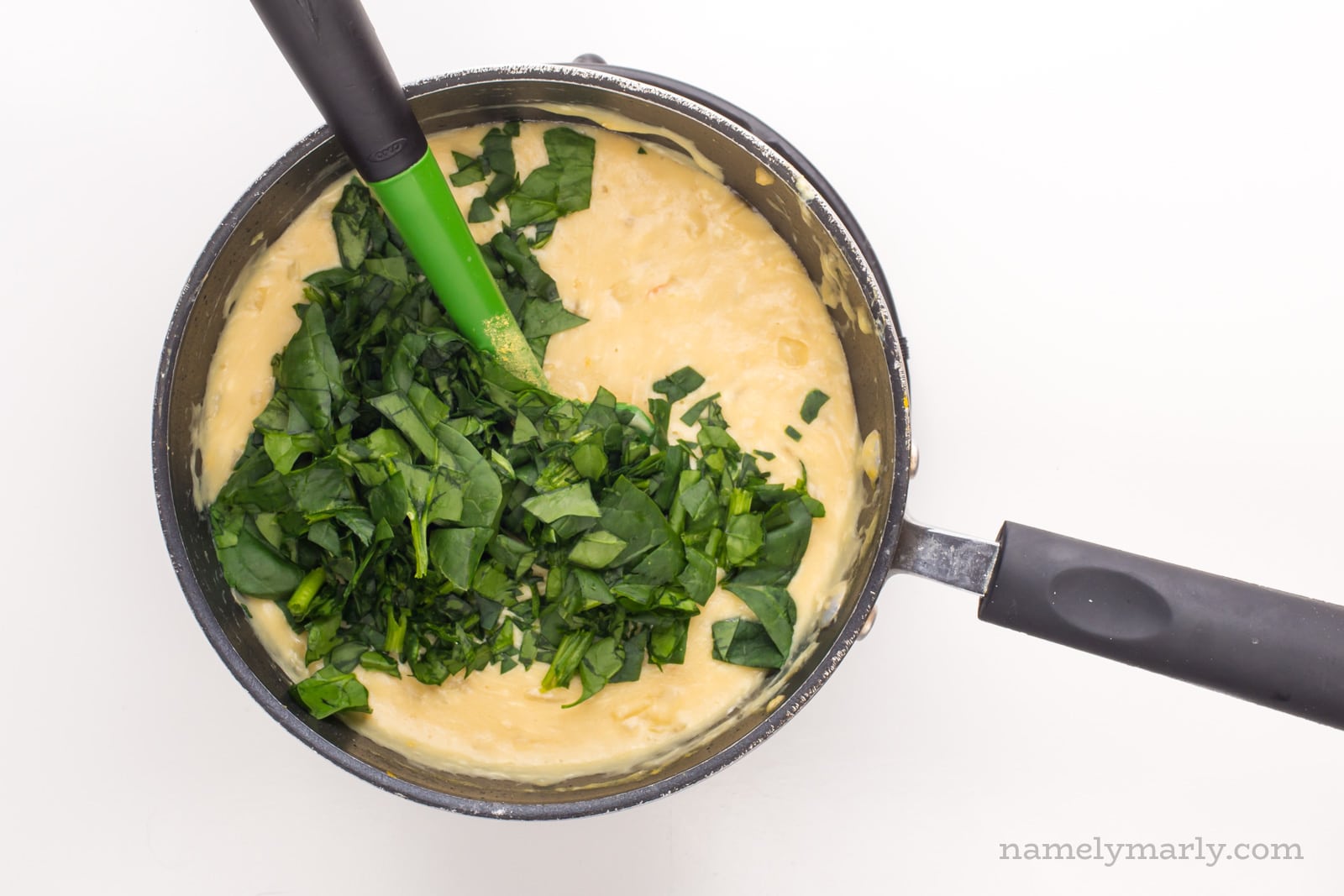 A saucepan shows the melted vegan cheese sauce with fresh spinach just added. A green spatula is in th epot, waiting to stir it all together.
