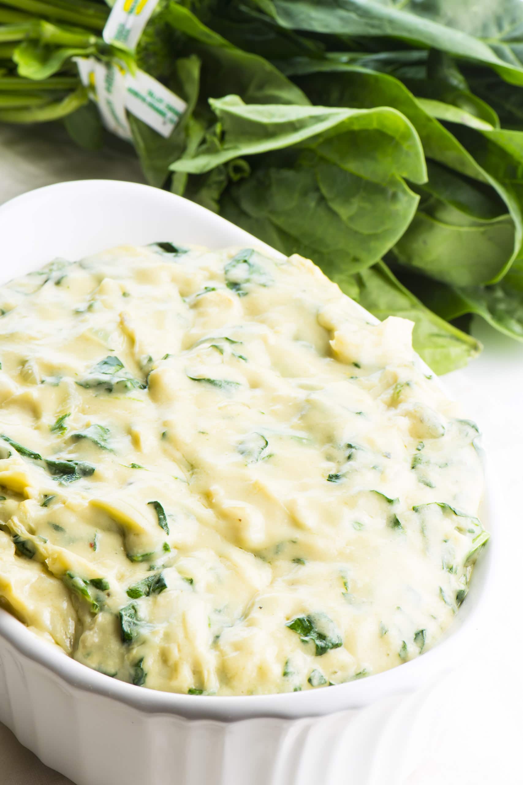 A white bowl with vegan spinach artichoke dip sits next to fresh spinach, an ingredient involved in the recipe.