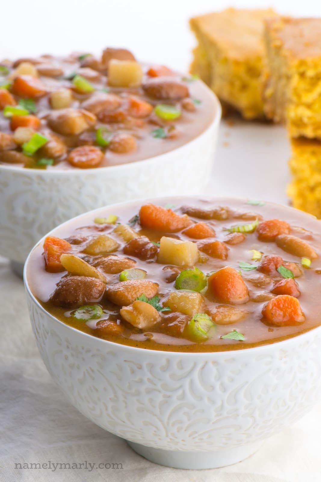 Two bowls of vegan pinto bean soup sit one in front of the other. The bowls are full of carrots, potatoes, green onions, and pinto beans. On the side are slices of cornbread.