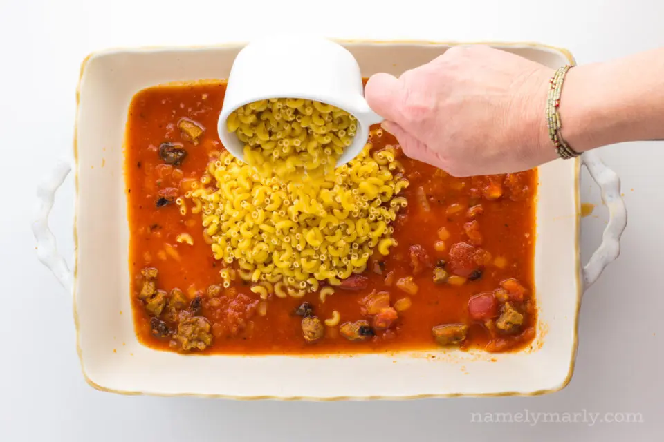 A hand holds a measuring cup pouring dry noodles over the sauce in the bottom of the casserole dish.