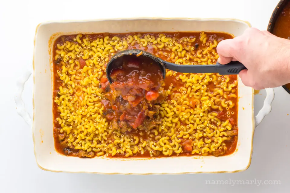 A hand holds a black spoon, pouring red sauce over noodles in a baking dish.