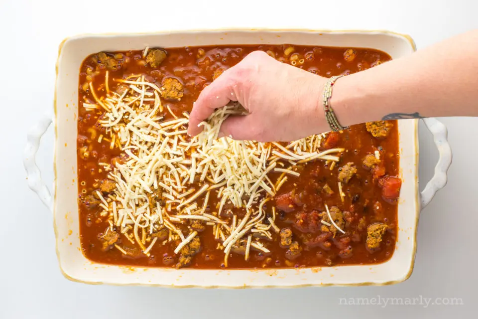 A hand is dropping vegan mozzarella shreds over a casserole dish.