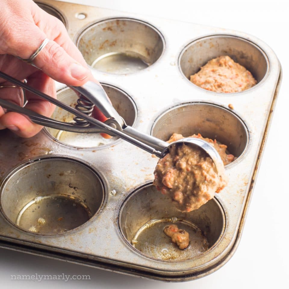 A hand reaches in with a cookie dispenser to add carrot cake muffin batter to an empty compartment of a muffin tin.