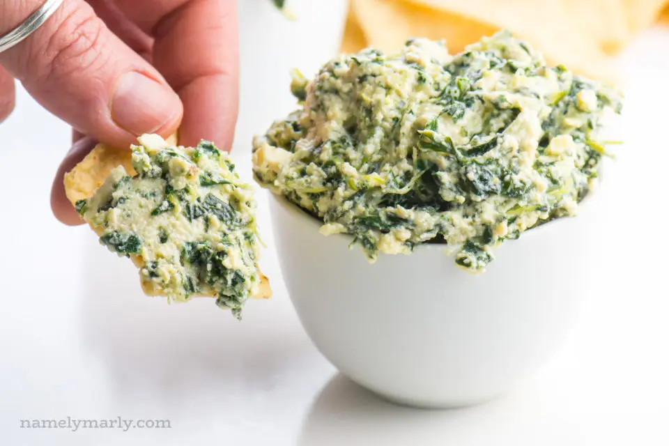 Vegan Spinach dip is served in a white container. A hand is holding a tortilla chip full of dip beside it. More tortilla chips are in the background.