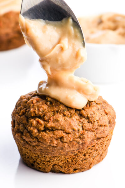 A spoon is dropping some tasty frosting on a vegan carrot cake muffin. There's a white dish with more frosting in the background and more muffins too.