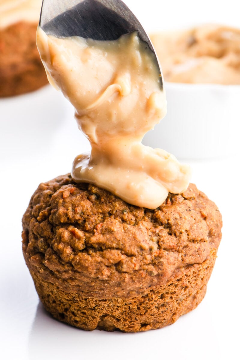 A spoon is dropping some tasty frosting on a vegan carrot cake muffin. There's a white dish with more frosting in the background and more muffins too.