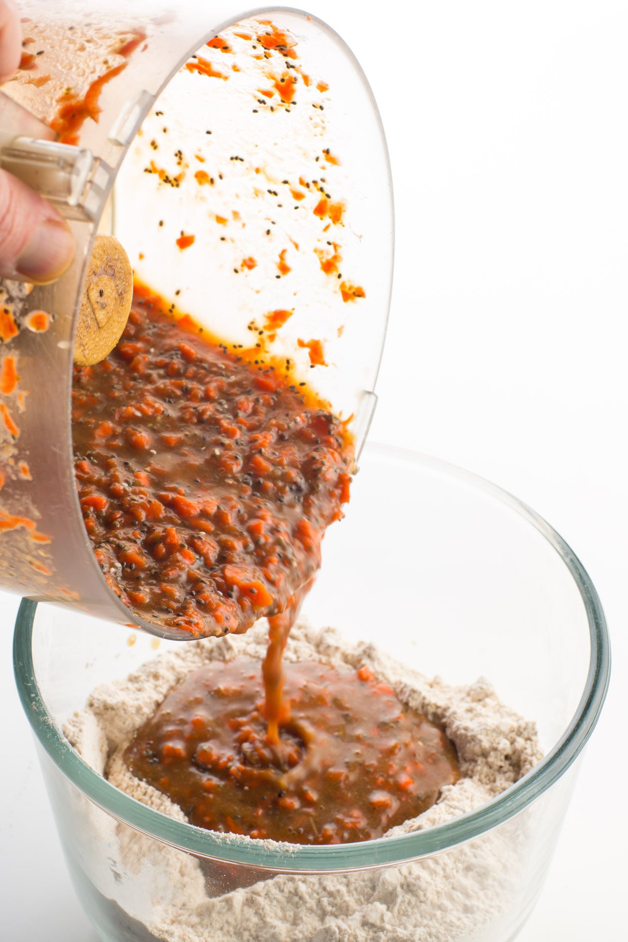 Ingredients from a food processor are being poured into a glass bowl full of the flour ingredients.