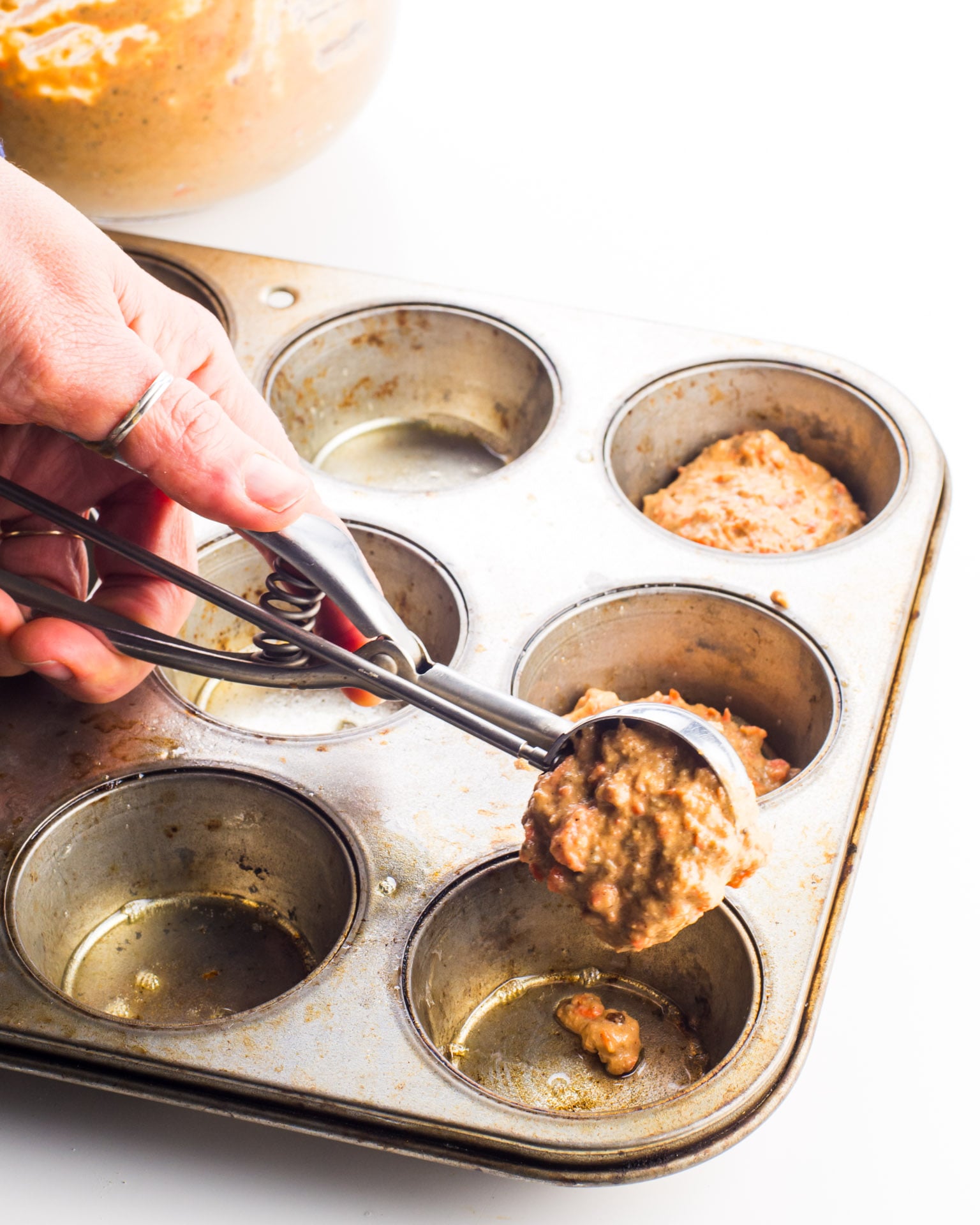 A hand reaches in with a cookie dispenser to add carrot cake muffin batter to an empty compartment of a muffin tin.