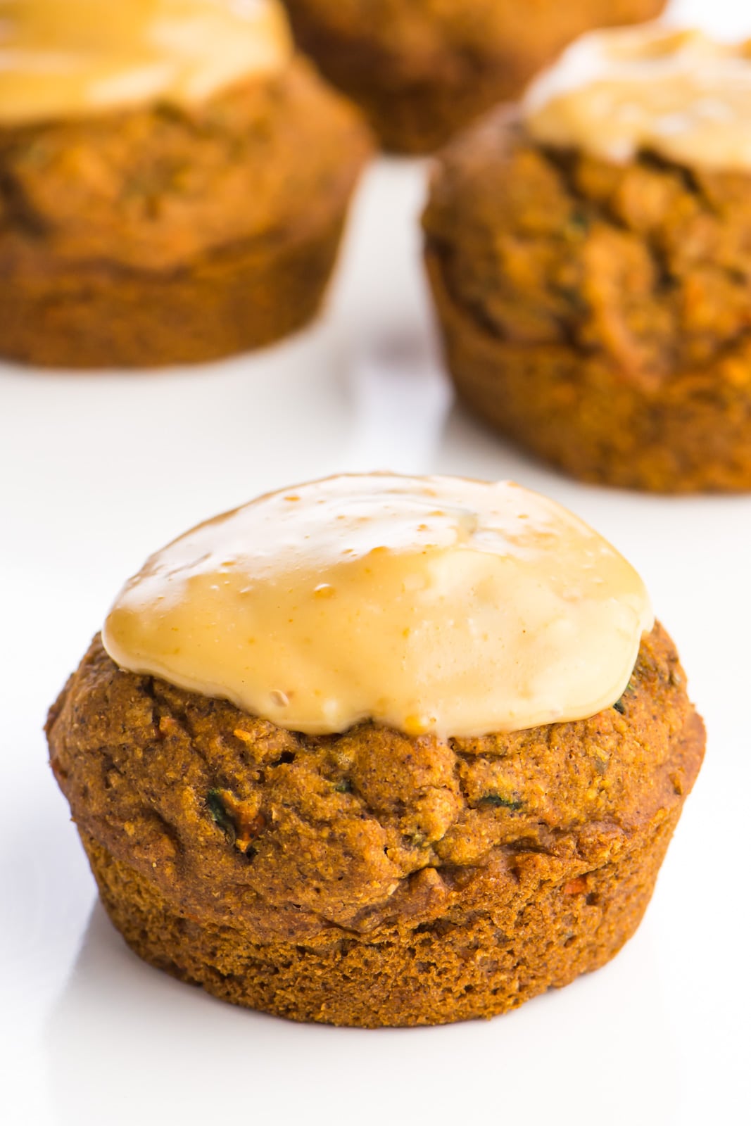 A vegan carrot cake muffin sits in the foreground with some maple drizzle on the top. There are more muffins int he background.