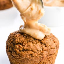 A spoon is dropping some tasty frosting on a vegan carrot cake muffin. There's a white dish with more frosting in the background and more muffins too.