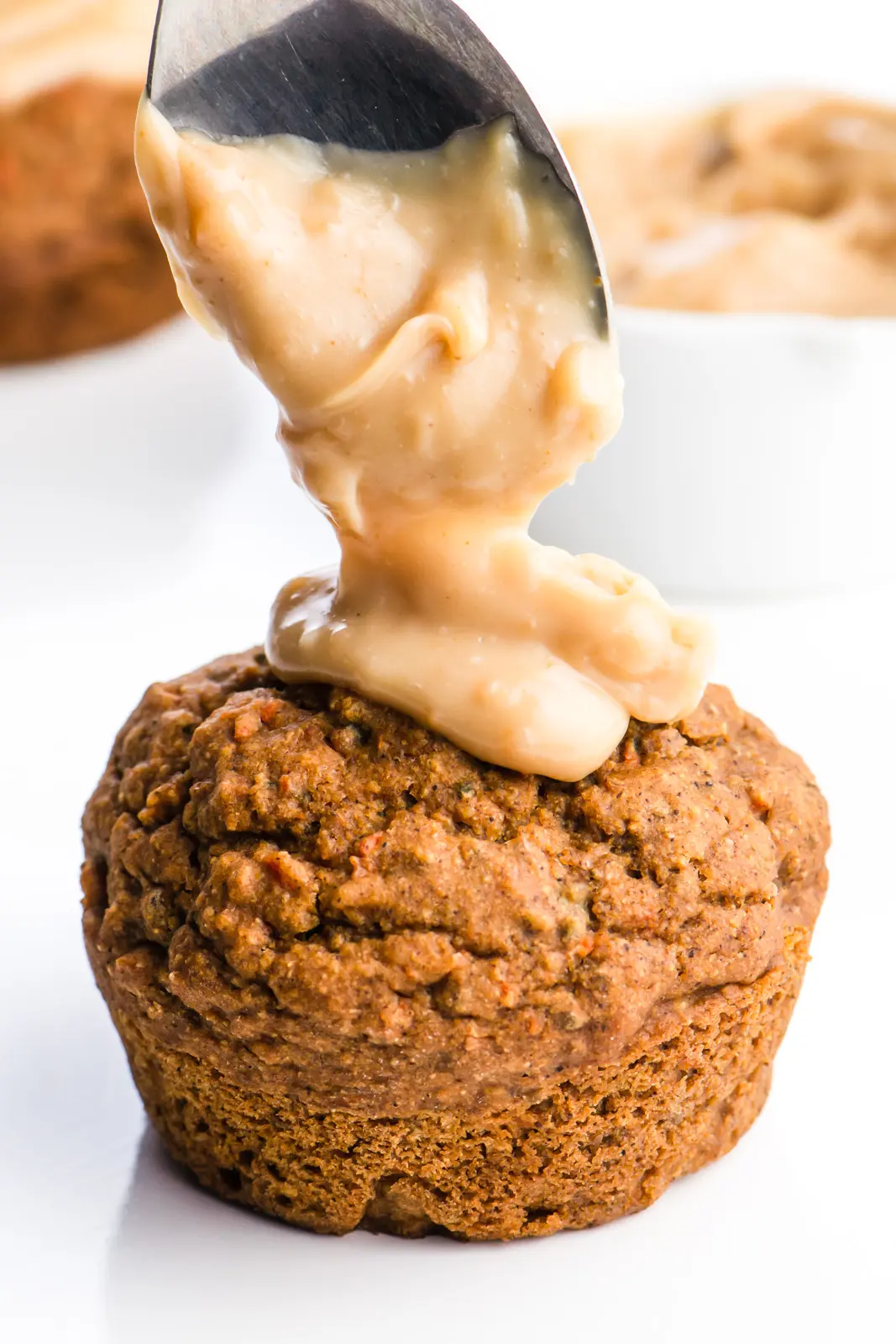  A spoon is dropping some tasty frosting on vegan carrot cake muffins. There's a white dish with more frosting in the background and more muffins too.